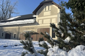 Jasny budynek z dwuspadowym dachem, częsciowo pokrytym śniegiem, widoczny na tle niebieskiego nieba. Przed budynkiem leży warstwa śniegu, a na pierwszym planie znajdują się gałęzie sosny z osadzonym śniegiem. Na elewacji widczony napis "Spichlerz Dworski"