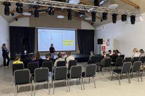Sala konferencyjna z ustawionymi w rzędach krzesłami skierowanymi w stronę sceny. Na scenie znajduje się ekran z wyświetlaną prezentacją oraz sprzęt nagłaśniający i oświetleniowy. 