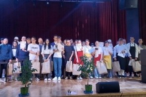 Grupa osób stoi na scenie, część z nich trzyma dyplomy, statuetki oraz papierowe torby. W tle widoczna jest bordowa kurtyna, a po bokach sceny ustawiono głośniki. Przed sceną znajdują się rośliny doniczkowe oraz elementy wyposażenia technicznego.