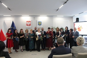 Grupa osób pozuje w sali sesyjnej. Uczestnicy trzymają róże i teczki, a w tle widoczne są flagi, godło Polski oraz herb Powiatu Cieszyńskiego.
