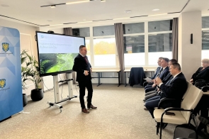 Mężczyzna prowadzi prezentację w sali konferencyjnej przed siedzącymi uczestnikami. Na ekranie widoczny jest slajd dotyczący programu redukcji ryzyka powodziowego, a obok znajduje się ścianka z herbem Powiatu Cieszyńskiego.