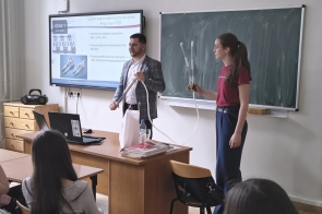 Sala wykładowa z tablicą kredową i ekranem prezentacyjnym. Mężczyzna i młoda dziewczyna stoją przy biurku; oboje trzymają elementy z przezroczystymi rurkami. Na ekranie wyświetlona jest prezentacja z ilustracjami urządzeń medycznych i tekstem. Przed nimi siedzą osoby skierowane w stronę prowadzących. Na biurku znajduje się laptop, butelka wody i stos dokumentów.