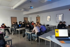 Uczniowie pracujący w grupach przy stolikach, na biurkach laptopy, z przodu otwarty komputer i projektor pod sufitem.