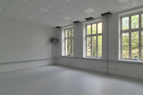 Wyremontowane pomieszczenie z jasnymi ścianami, dużymi oknami i nową podłogą. Przy ścianach widoczne są elementy instalacji, a w suficie znajdują się częściowo otwarte panele.