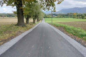 Wyremontowana droga asfaltowa prowadzi przez pola i tereny zielone. Po lewej stronie widoczny jest szpaler drzew, a w tle rozciąga się widok na zabudowania i pasmo górskie.