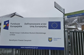 Tablica informacyjna o dofinansowaniu z Unii Europejskiej ustawiona przy ogrodzeniu, obok znak kierunkowy i zabudowania na tle ośnieżonego zbocza.