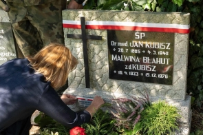 Kobieta przy grobie ozdobionym biało-czerwoną wstęgą umieszcza oznaczenie na płycie nagrobnej. Wokół widoczne są rośliny oraz kwiaty.