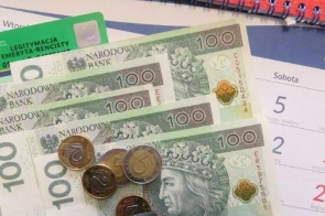 Kilka banknotów o nominale 100, ułożonych w wachlarz, oraz monety w różnych wartościach leżą na papierowej powierzchni. W tle widoczny jest fragment kalendarza z zaznaczonymi sobotami i niedzielami oraz plastikowa karta w zielonym kolorze z białym napisem, sugerująca dokument potwierdzający status emeryta lub rencisty. Spiralny grzbiet kalendarza dodaje kompozycji biurowego charakteru.