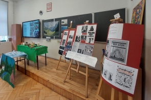 Sala lekcyjna z podwyższeniem przy tablicy. Na sztalugach ustawione są plansze z tekstami i zdjęciami, w tym reprodukcjami okładek książek i fotografiami czarno-białymi. Z boku stoi stół przykryty zielonym obrusem z makietami domów. Na ścianie widoczna jest tablica, ekran z wyświetloną prezentacją, zegar oraz godło Polski.