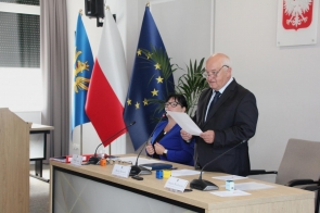Maciej Bieniek Przewodniczący Rady w ciemnym garniturze stoi przy stole i trzyma kartkę z dokumentem. Obok niego siedzi Klaudia Kaleta Sekretarz ubrana w niebieską marynarkę. W pomieszczeniu znajdują się mikrofony, dokumenty oraz tabliczki z nazwiskami, a w tle widoczne są flagi.