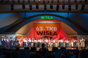 Grupa osób w strojach regionalnych stoi w rzędzie na scenie. Po lewej stronie widoczni są muzycy z instrumentami, a po prawej dwie kobiety stoją przy mikrofonach. W tle znajduje się napis „63. TKB Wisła”. Oświetlenie sali jest ciemne, a światło sceniczne pada głównie na stojące osoby.