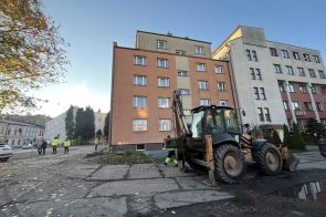 Na placu przed budynkiem trwa remont. Widoczna jest koparko-ładowarka, pracownicy w odzieży odblaskowej oraz fragment zabezpieczenia terenu robót. W tle znajduje się kilkupiętrowy budynek i sąsiednia zabudowa.