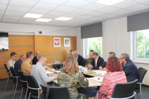 Kilkanaście osób siedzi przy okrągłym stole konferencyjnym i uczestniczy w spotkaniu. Na stole znajdują się dokumenty, notatki, filiżanki, butelki z wodą oraz sprzęt multimedialny. Wśród uczestników widoczne są osoby w stroju cywilnym oraz mundurach. Spotkanie odbywa się w jasnej sali z dużymi oknami.