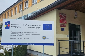 Budynek szkoły o żółto-pomarańczowej elewacji z wejściem i podjazdem. Przed nim stoi tablica informacyjna o dofinansowaniu z Funduszy Europejskich.