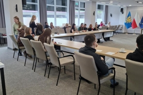 Sala sesyjna z ustawionymi w kształt litery U stołami i krzesłami. Przy stołach siedzi kilkunastu uczniów, część stoi. W tle widoczne są okna oraz ściana z flagami i logo na jasnym tle.