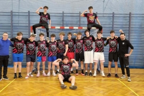 Hala sportowa z drewnianą podłogą i bramką w tle. Grupa nastolatków w czarno-czerwonych koszulkach z grafiką niedźwiedzia pozuje do zdjęcia razem z dwoma dorosłymi mężczyznami stojącymi po bokach. Dwóch chłopaków siedzi na górnej belce bramki, a jeden siedzi na podłodze z przodu i pokazuje zgięte ręce.