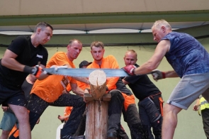 Pięciu mężczyzn przecina drewnianą belkę dużą piłą ręczną. Trzech z nich ma pomarańczowe koszulki, a pozostali czarne i granatowe ubrania. W tle stoją kolejne osoby.