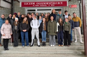 Wejście do budynku z kamienną elewacją i czerwonym szyldem „I Liceum Ogólnokształcące im. Pawła Stalmacha w Wiśle”. Na schodach stoi duża grupa młodzieży oraz kilka dorosłych osób, ubranych w kurtki i bluzy. Wszyscy pozują do wspólnego zdjęcia przed drzwiami szkoły.