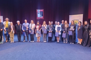 Grupa laureatów i organizatorów stojących na scenie teatru, trzymających dyplomy i nagrody, na tle kurtyny i banerów powiatu cieszyńskiego.