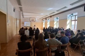 Zdjęcie przedstawia salę szkolną z grupą uczniów siedzących na krzesłach ustawionych w rzędach. Uczniowie są zwróceni w stronę ekranu, na którym wyświetlana jest prezentacja. Po lewej stronie widoczne okna, na ścianach dekoracje.