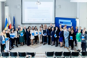 Grupa kilkudziesięciu osób pozuje do wspólnego zdjęcia w jasnej sali konferencyjnej, wiele z nich trzyma niebieskie teczki. W tle widoczne są flagi, ekran z prezentacją o Funduszach Europejskich oraz baner informacyjny.