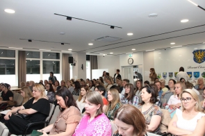Duża grupa osób siedzi w rzędach w jasnej sali konferencyjnej. Większość uczestników jest zwrócona w jednym kierunku. W tle widoczne są herby gmin oraz herb Powiatu Cieszyńskiego.
