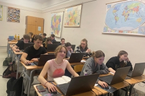 Uczniowie siedzą w sali lekcyjnej przy biurkach z laptopami, skupieni na pracy. Na ścianach wiszą mapy świata i Afryki. W tle stoją butelki z napojami. Jedna z uczennic uśmiecha się do obiektywu.