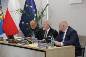 Trzech mężczyzn siedzi za stołem prezydialnym z mikrofonami, dokumentami i butelkami wody. W tle widać flagi, roślinę oraz jasną salę obrad.