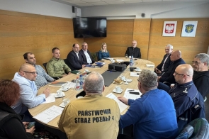 Grupa osób siedzących przy stole konferencyjnym podczas spotkania, z dokumentami, kawą i wodą, w tle ściana z herbami i ekranem.