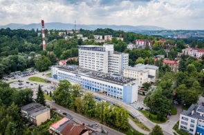 Rozległy kompleks budynków otoczony zielenią, widziany z lotu ptaka. Wokół znajdują się drogi, parkingi i zabudowa miejska, a w tle widoczne są wzgórza i góry.