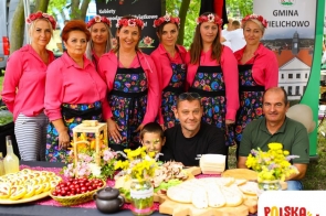 Grupa kobiet w różowych koszulach i kwiecistych fartuchach, z wiankami na głowach, stoi za stołem pełnym tradycyjnych potraw. Na stole widać chleb, wędliny, wiśnie i kwiaty. Obok nich siedzi dwóch mężczyzn i chłopiec. Scena odbywa się na tle zielonego parku.