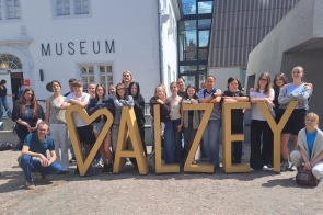 Grupa młodzieży i opiekunów pozuje przed budynkiem muzeum. Uczestnicy stoją wokół dużej przestrzennej instalacji tworzącej napis „ALZEY”, ustawionej na brukowanym placu. W tle widoczna jest jasna elewacja oraz nowoczesna zabudowa z dużymi przeszkleniami.