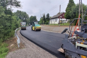 Maszyny drogowe układają i zagęszczają nową warstwę asfaltu na jezdni. Wzdłuż drogi widoczne są bariery ochronne, chodnik oraz zabudowania otoczone zielenią.