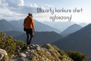 Grafika przedstawia osobę w pomarańczowej kurtce i czarnych spodniach stojącą na skalistym szczycie, patrzącą na widoczne w oddali pasma górskie. Na jasnym niebie ponad szczytami gór widnieje czarny napis „Otwarty konkurs ofert ogłoszony!”.