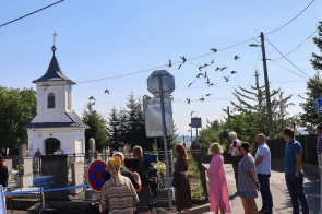 Grupa osób stoi przy ogrodzeniu w pobliżu niewielkiej kaplicy. Nad drogą przelatuje stado ptaków, a obok ustawiony jest sprzęt filmowy i znaki drogowe.
