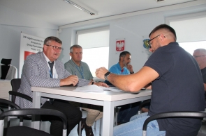 Na zdjęciu widać grupę mężczyzn siedzących przy stole w biurze. Dwóch z nich siedzi po prawej stronie stołu, a trzech po lewej Jerzy Pilch, Członek Zarządu, Jarosław Kalinik, Dyrektor Powiatowego Zarządu Dróg Publicznych w Cieszynie, Ryszard Krzywoń, Zastępca Dyrektora Powiatowego Zarządu Dróg Publicznych w Cieszynie. Pierwszy mężczyzna po lewej stronie: Jerzy Pilch ma na sobie marynarkę w kratkę i okulary, trzyma długopis i patrzy na dokumenty leżące na stole. Drugi mężczyzna: Jarosław Kalinik siedzący obok niego ma na sobie szarą koszulę z długim rękawem. Trzeci mężczyzna: Ryszard Krzywoń ma na sobie niebieską koszulę. Po prawej stronie stołu siedzi mężczyzna w ciemnej koszulce z krótkim rękawem, również z okularami. Za nim widać kolejnego mężczyznę w okularach. Na ścianie wiszą dwa godła oraz tablica z napisem.