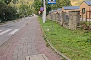 Na zdjęciu widoczny jest fragment ulicy z chodnikiem wykonanym z kostki brukowej oraz przejściem dla pieszych. Wzdłuż drogi znajdują się znaki drogowe, pas zieleni oraz ogrodzenie z kamiennymi słupkami. Po prawej stronie widoczne są budynki o jasnych elewacjach i ciemnych dachach.