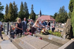 Grupa osób stoi przy grobie na cmentarzu. Dwie osoby na pierwszym planie układają kwiaty i znicze, a pozostali stoją obok. W tle widoczne są nagrobki, drzewa i budynek z wieżyczką.