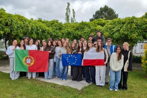 Grupa młodzieży pozuje do wspólnego zdjęcia z flagami Portugalii, Polski i Unii Europejskiej. Zielone otoczenie oraz pochmurne niebo.