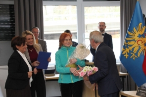 Grupa osób stoi w sali sesyjnej podczas uroczystości. Jedna osoba trzyma zapakowany upominek, a druga podaje bukiet kwiatów. W tle widoczna jest flaga Powiatu Cieszyńskiego oraz okna z zasłonami.