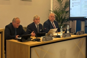 Po lewej stronie siedzi Grzegorz Konieczny Wiceprzewodniczący Rady ubrany w czarny garnitur, obok niego Stanisław Kubicius Przewodniczący Rady w granatowym garniturze, a następnie Jarosław Bodak Wiceprzewodniczący Rady w niebieskim garniturze. Przed nimi stoją tabliczki z imionami i nazwiskami, tablety oraz laptop, a także butelki z wodą i szklanki. Po prawej stronie widoczny jest ekran z wyświetlonym porządkiem obrad, w tle znajduje się roślina doniczkowa.