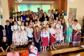 Do wspólnego zdjęcia pozują dzieci, młodzież oraz osoby dorosłe. Część uczestników ubrana jest w stroje regionalne. Grupa ustawiona jest na scenie na tle dekoracji przedstawiającej krajobraz.