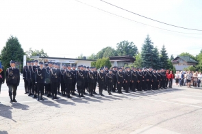 Duża grupa strażaków w mundurach galowych stoi w równych szeregach na placu. Po prawej stronie za ogrodzeniem stoją osoby obserwujące uroczystość, a w tle widoczne są budynki i drzewa.