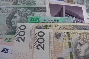 Rozłożone są banknoty o nominałach 100 i 200 złotych, a pomiędzy nimi leżą dokumenty i karty z logo ZUS, w tym zielona legitymacja emeryta-rencisty. Kolory tworzą kontrast zieleni, bieli i beżu, a w tle widać fragment fotografii budynku.