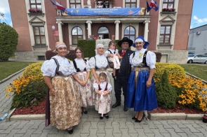 Grupa osób w strojach regionalnych pozuje przed budynkiem udekorowanym na dożynki. W tle widoczne są flagi, baner wydarzenia oraz rabaty z żółtymi kwiatami.