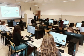 Sala komputerowa, w której uczniowie siedzą przy biurkach i pracują na komputerach. Na monitorach otwarty jest program do projektowania 3D, a na ścianie wyświetlony jest obraz z projektora z tym samym programem. Przy tablicy stoi nauczyciel.
