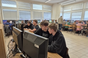 Uczniowie siedzą przy stanowiskach komputerowych w pracowni i pracują przy monitorach. Część osób siedzi parami, a w tle widoczne są kolejne stanowiska oraz osoba stojąca między rzędami komputerów.