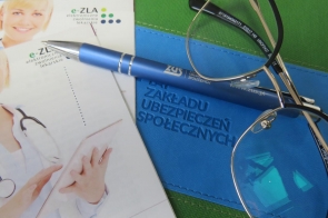 Na zdjęciu widać teczkę oraz dwie ulotki na których znajduję się postać lekarki w młodym wieku. Obok leżą okulary wraz z niebieskim długopisem z logiem ZUS.