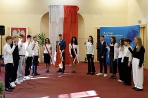Grupa uczniów w strojach galowych stoi w sali z polskimi flagami, przygotowana do występu lub apelu.