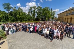 Uczestnicy wydarzenia pozują do wspólnego zdjęcia na dziedzińcu przed budynkiem. Na fotografii widoczna jest liczna grupa osób zgromadzonych na otwartej przestrzeni. W tle znajdują się drzewa, park oraz zabudowania.